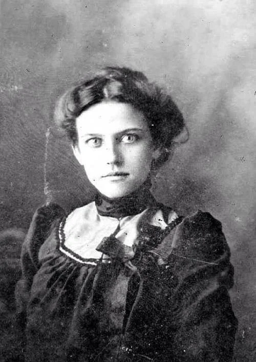 Black and white portrait of a woman from the early 1900s, with wavy hair and wearing a high-collared, ruffled dress. She looks directly at the camera with a neutral expression. The background is blurred.