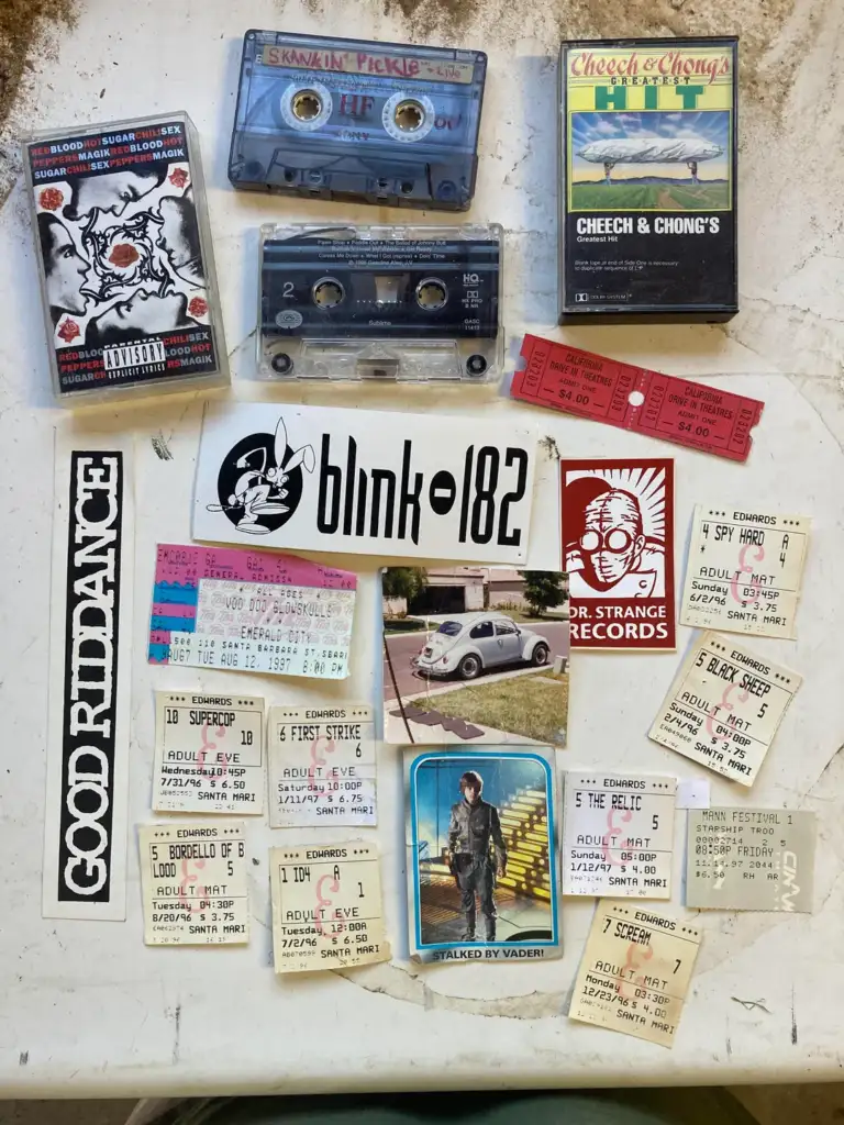 A collection of vintage concert tickets, cassette tapes, and stickers arranged on a white surface. Featured are band tickets, a Blink-182 sticker, and various cassette covers, including a skull design and a classic car image.