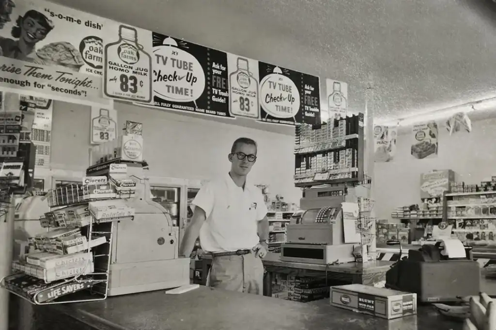 A black-and-white photo of a man standing in a retro grocery store. The store is filled with various products, including chewing gum and household items. Signs above advertise items like Clorox and suggest "TV tube check-up time." The man wears glasses and a white shirt.