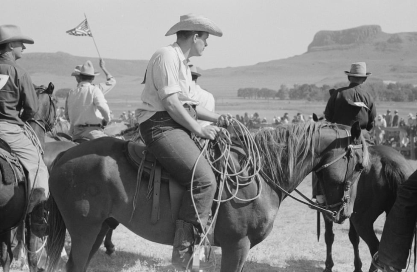 A cowboy wearing a hat and jeans sits on a horse in a grassy field. Other riders and horses are gathered in the background. One rider holds an American flag. A mesa is visible in the distance under a clear sky.