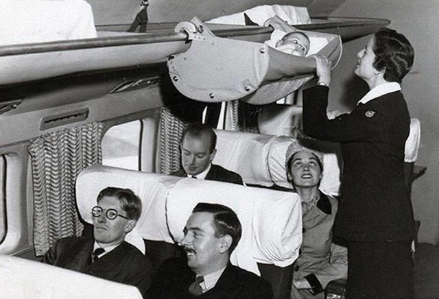 Vintage photo of an airplane cabin featuring an overhead storage area with a suspended baby cradle. A flight attendant stands nearby, ensuring the baby is secure. Passengers are seated below, observing the scene with interest.