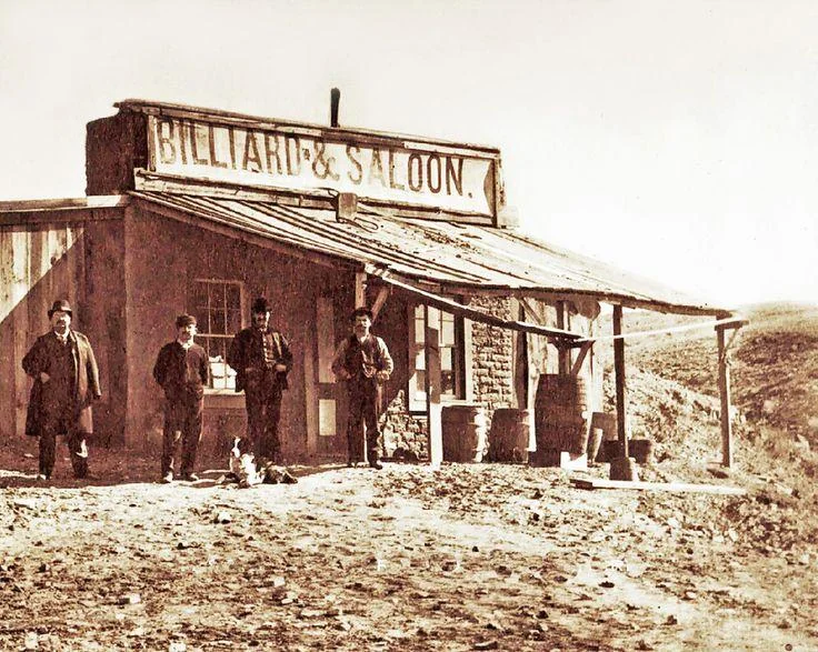 Four men stand outside a rustic, weathered building labeled "Billiard & Saloon." The scene depicts a barren landscape, capturing an old Western atmosphere with wooden barrels and a dirt foreground.