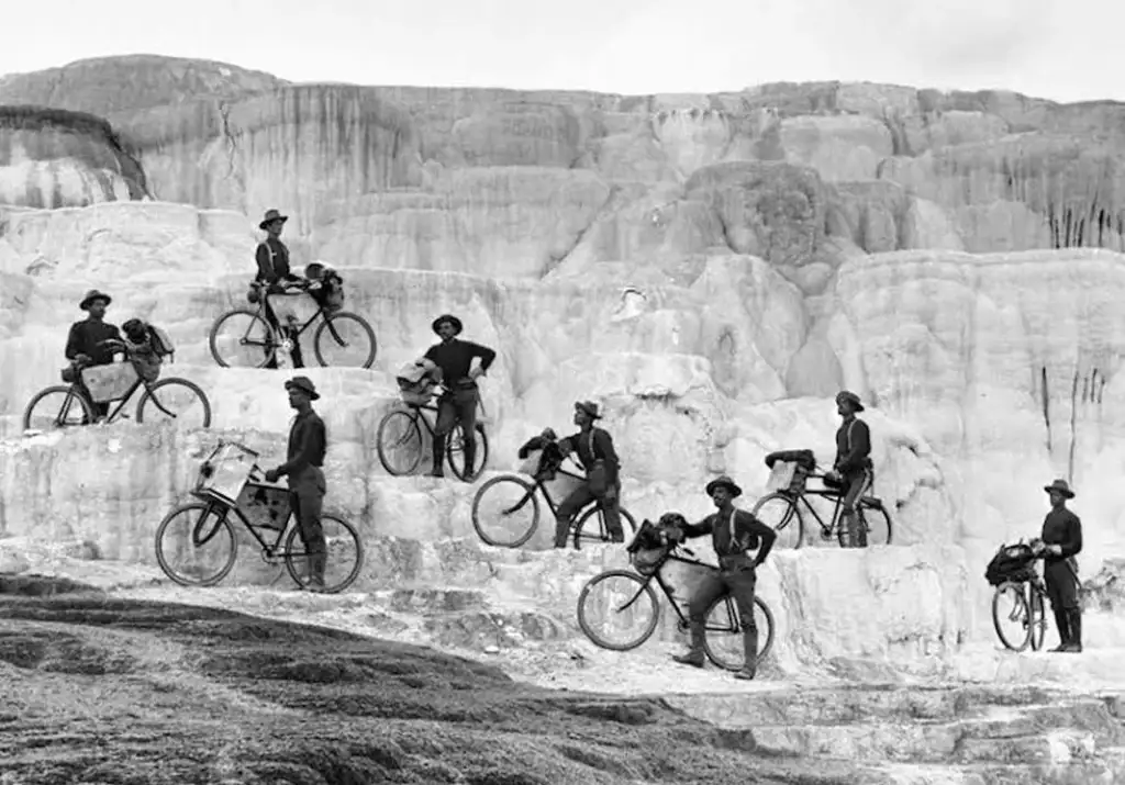 A group of nine soldiers in uniform poses with bicycles on the uneven, terraced landscape of Mammoth Hot Springs in Yellowstone National Park. The terraces have a stepped appearance with varying shades of gray and white.