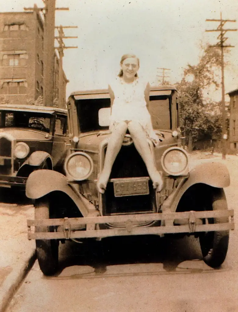 A vintage photo shows a woman in a dress smiling and sitting on the hood of an old car, parked on a street lined with power poles and buildings. Another vintage car is parked nearby. The scene evokes a nostalgic, early 20th-century feel.