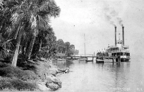 A vintage steamboat docked at a riverbank surrounded by palm trees, with smoke rising from its twin chimneys. A few small boats are nearby, and a bridge is visible in the background. The setting appears calm and serene.