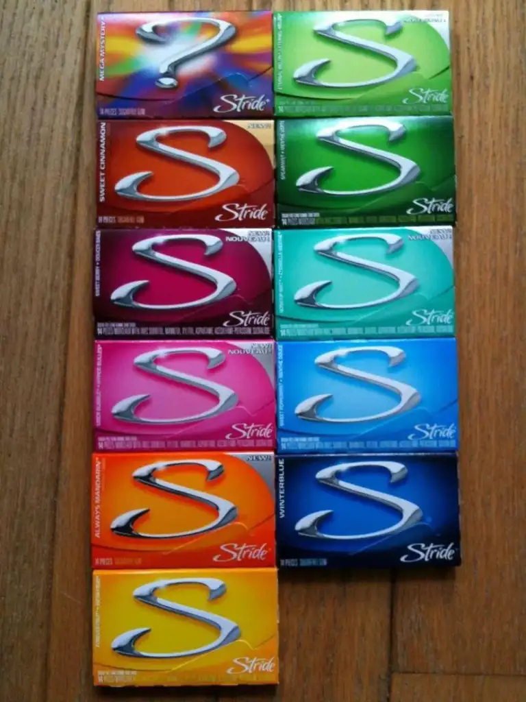 Ten packs of Stride gum in various colors are arranged on a wooden surface. Each pack has a large silver "S" logo and distinct colors, including shades of blue, green, pink, orange, and purple.