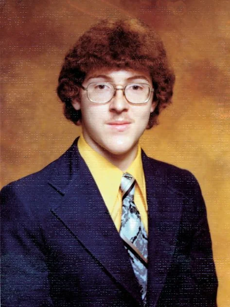 A person with curly hair and glasses is wearing a dark suit, yellow shirt, and patterned tie. The background is a warm brown gradient.