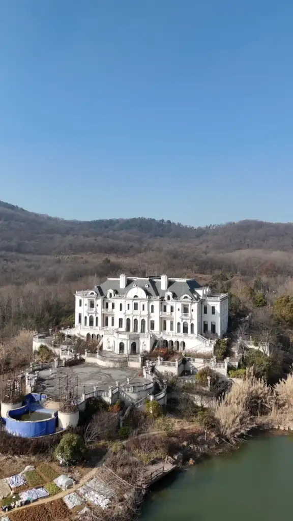 A large, white mansion with multiple chimneys stands in a forested area. The surrounding landscape has barren trees and hills under a clear blue sky. A circular blue structure is visible in the foreground near the lake.