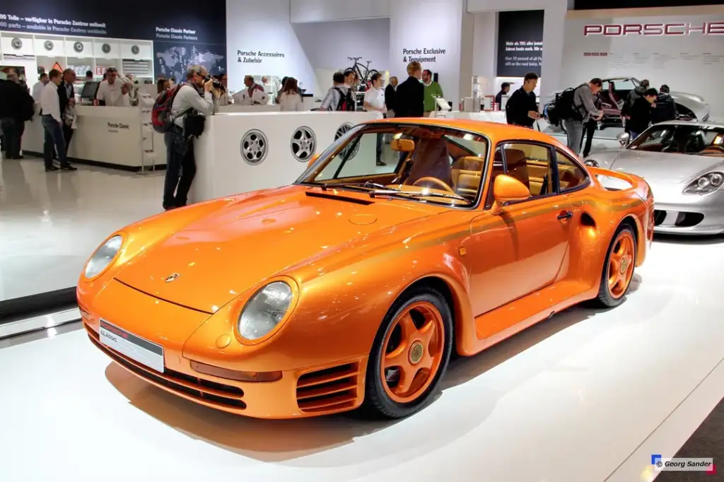 An orange Porsche 959 displayed at an auto show. The sleek sports car features distinctive curves and design, with people and other cars visible in the background, creating an atmosphere of a high-end car exhibition.