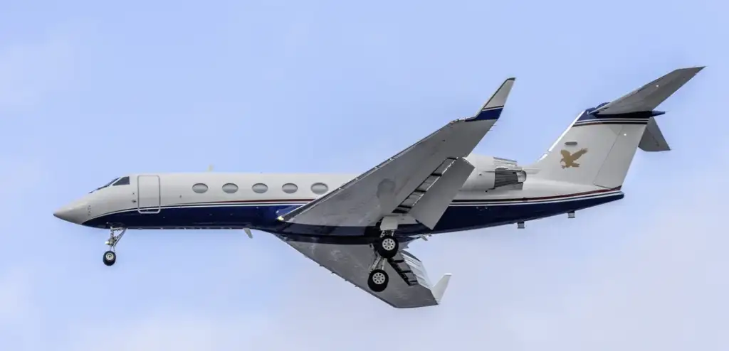 A sleek private jet with a white body and blue accents is flying against a clear blue sky. Its landing gear is extended, and the tail features a gold emblem of an eagle.