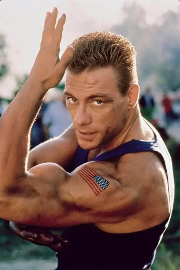 A muscular man in a sleeveless shirt poses with his arm flexed, showing an American flag tattoo on his shoulder. He has short hair and looks directly at the camera with a determined expression. The background is blurred outdoors.