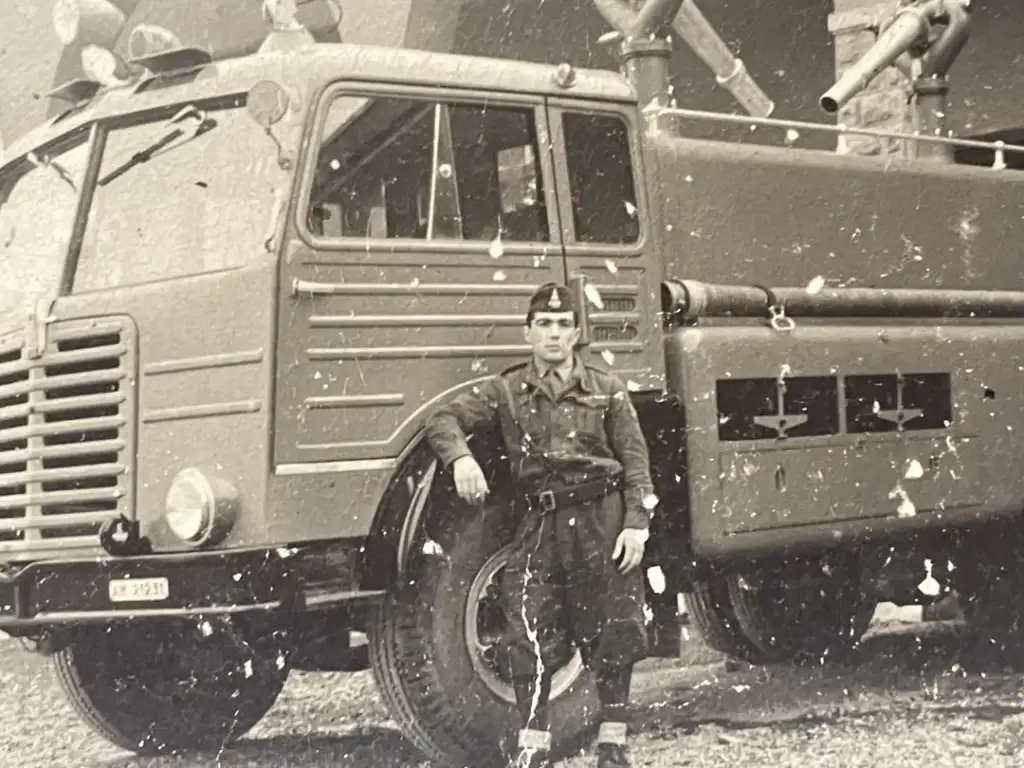 Black and white photo of a uniformed man standing beside a vintage fire truck. The truck has a ladder on top and various hoses and equipment. The man poses with one arm leaning on the front tire, and the surroundings appear to be outdoors.