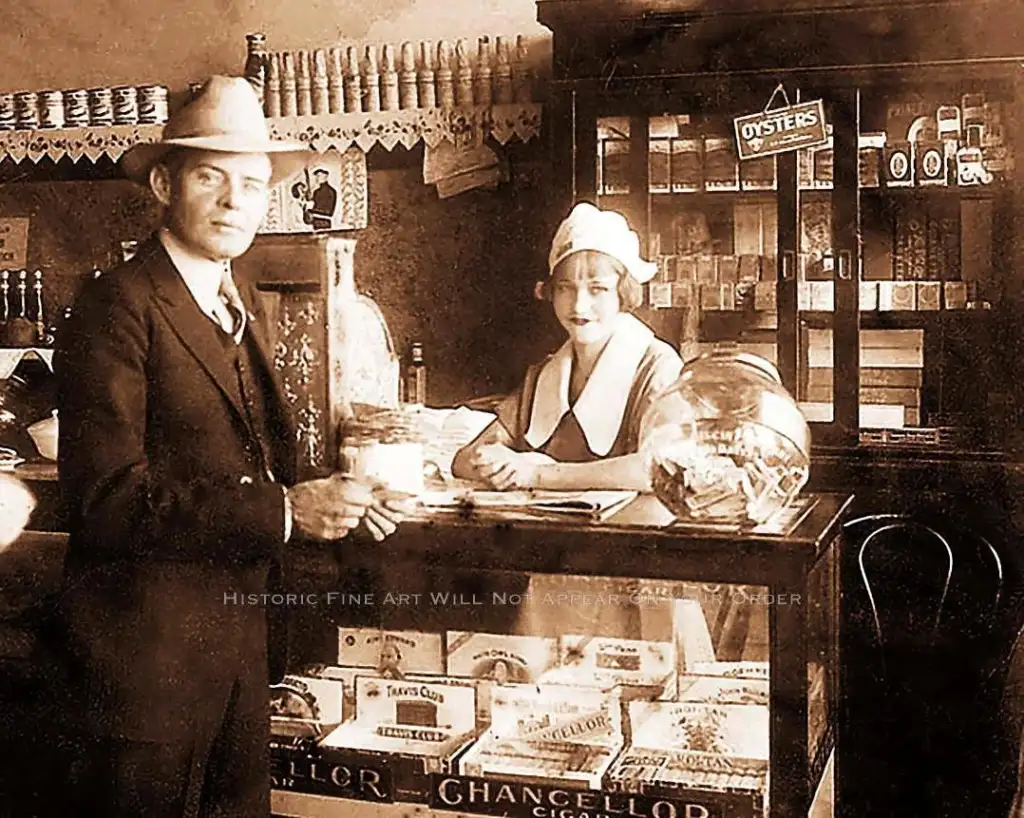 A vintage photo of a man and woman in a store. The man, in a suit and hat, stands by a counter with cigar boxes. The woman, in a uniform and hat, stands behind the counter. Shelves with groceries and a sign for oysters are in the background.