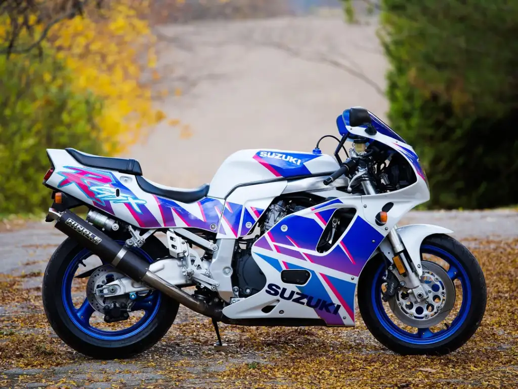 A white and blue Suzuki sport motorcycle parked on a leaf-covered road. The bike features vibrant purple and pink graphics. In the background, there are trees with green and yellow leaves.