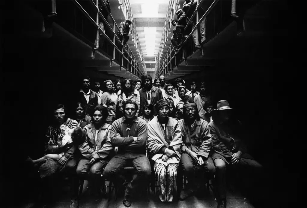 A black-and-white photo showing a large group of people seated and standing in rows within a dimly lit corridor. The ceiling has several skylights, illuminating the scene from above. The group looks directly at the camera.