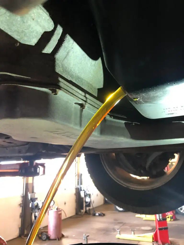 Engine oil draining from a vehicle's undercarriage, with a stream of golden liquid flowing into a container. The scene is inside an auto repair shop, visible with tools and equipment in the background.