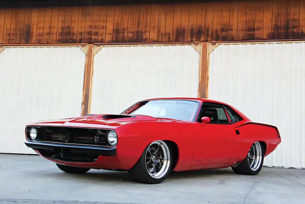 A classic red muscle car with sleek bodywork is parked on a concrete surface. It has a sporty, low profile, chrome rims, and a black hood scoop. The background features a wooden building with white garage doors.