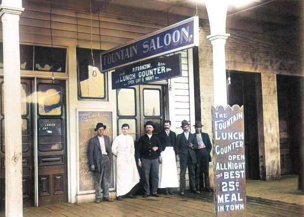 A group of men in vintage clothing stand outside a historical saloon with signs reading "Fountain Saloon" and "Lunch Counter open all night." Another sign advertises a 25-cent meal. The building has a wooden facade and pillars.