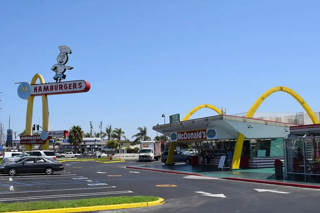 A retro-style McDonald's with vintage signage, including a large yellow arch and a classic "Hamburgers" sign featuring a chef figure. The parking lot is partially visible with a few cars parked under a clear blue sky.