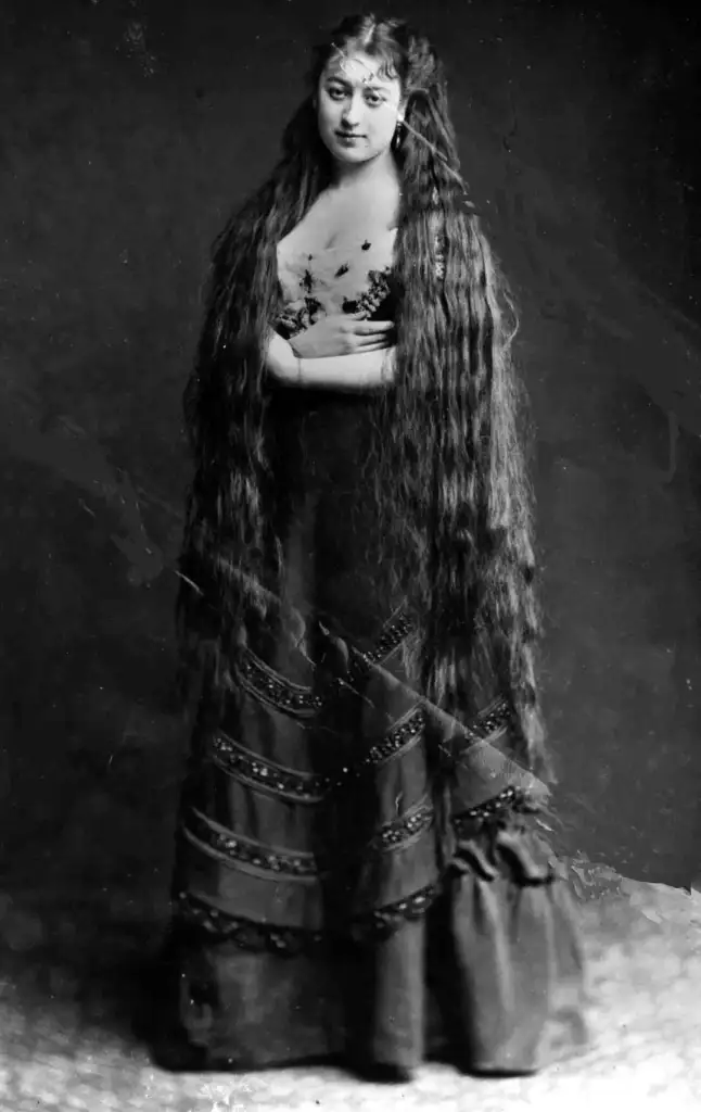 A woman stands with her arms crossed, wearing a long, dark dress adorned with decorative trims. Her very long hair flows down to her skirt. The photograph is black and white, giving it a vintage appearance.