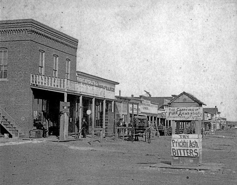 Black and white photo of a historical street scene with multiple wooden storefronts. A large sign in the foreground reads "Try Prickly Ash Bitters." A few people stand near the buildings, and a horse-drawn cart is visible.