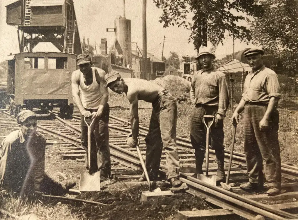 Five men are working on a railway track. Four are standing, holding shovels, and one is crouching. They are wearing work clothes and caps. A train and industrial buildings are visible in the background. The photo is in black and white.