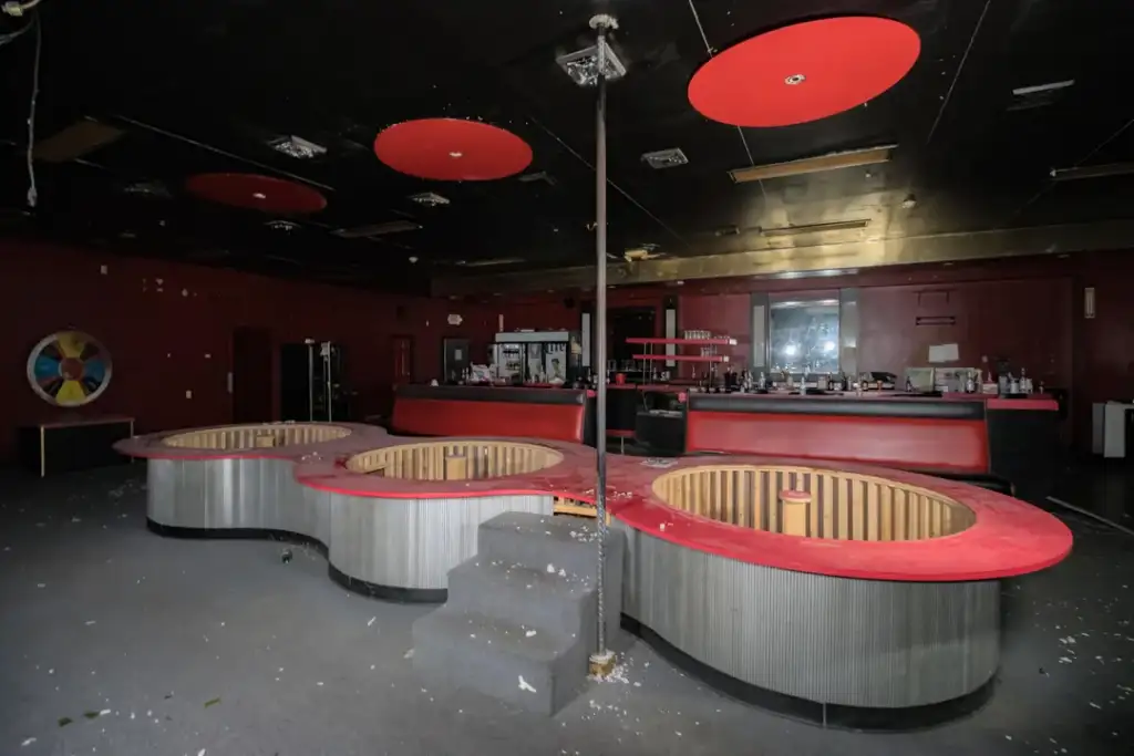 An abandoned nightclub interior with a dark red and black color scheme. Three circular, sunken seating areas with red rims are prominent. A pole stands in the center, and a bar with mirrored backing is visible in the background. Debris is scattered on the floor.