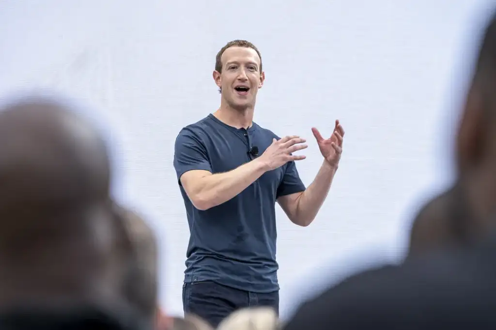 A man in a dark t-shirt is speaking on stage with expressive hand gestures. The background is a pale, solid color, and there are blurred heads in the foreground, indicating an audience.