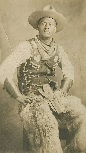 Vintage photo of a man dressed in Western cowboy attire, including a hat, leather vest with decorative patterns, scarf, and chaps. He is seated with hands on hips, exuding a confident and stoic expression.