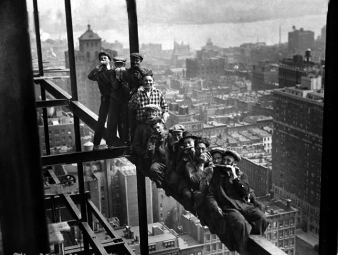 Black and white photo of eleven construction workers sitting on a steel beam high above a city skyline, eating lunch and posing for the camera, with buildings visible in the background.