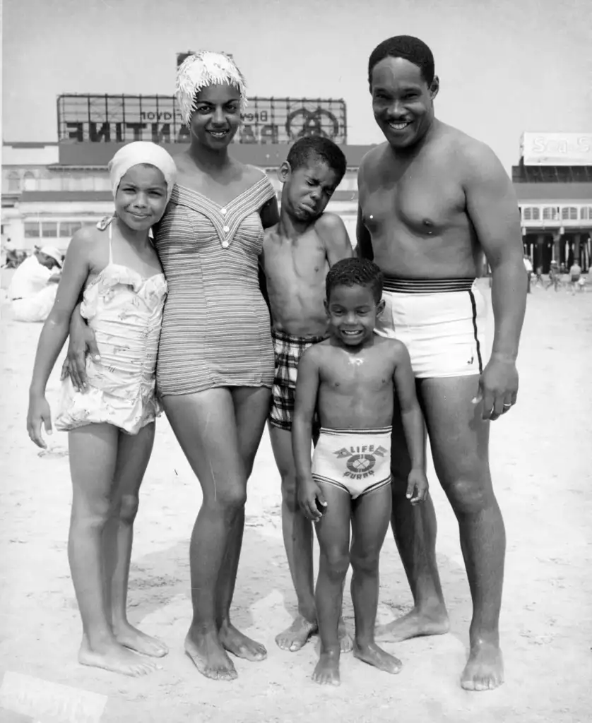 A family of five poses on a beach with a boardwalk in the background. Two adults stand with three children, all smiling. The girls wear swimsuits, and the boys wear swim trunks. Signs and beachgoers are visible in the distance.