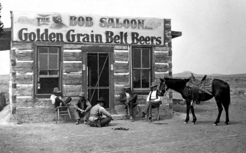 A black-and-white image of a rustic wooden saloon with a sign reading "Golden Grain Belt Beers." Four men sit and stand outside. A saddled horse is tied nearby. The scene evokes an Old West atmosphere.