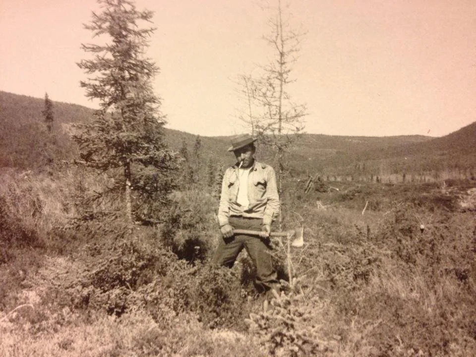 A man wearing a hat and work clothes stands in a rugged, sparsely wooded landscape with small trees and bushes. He's holding an object in one hand and stands near a young tree, with hills visible in the background. The photo is in black and white.