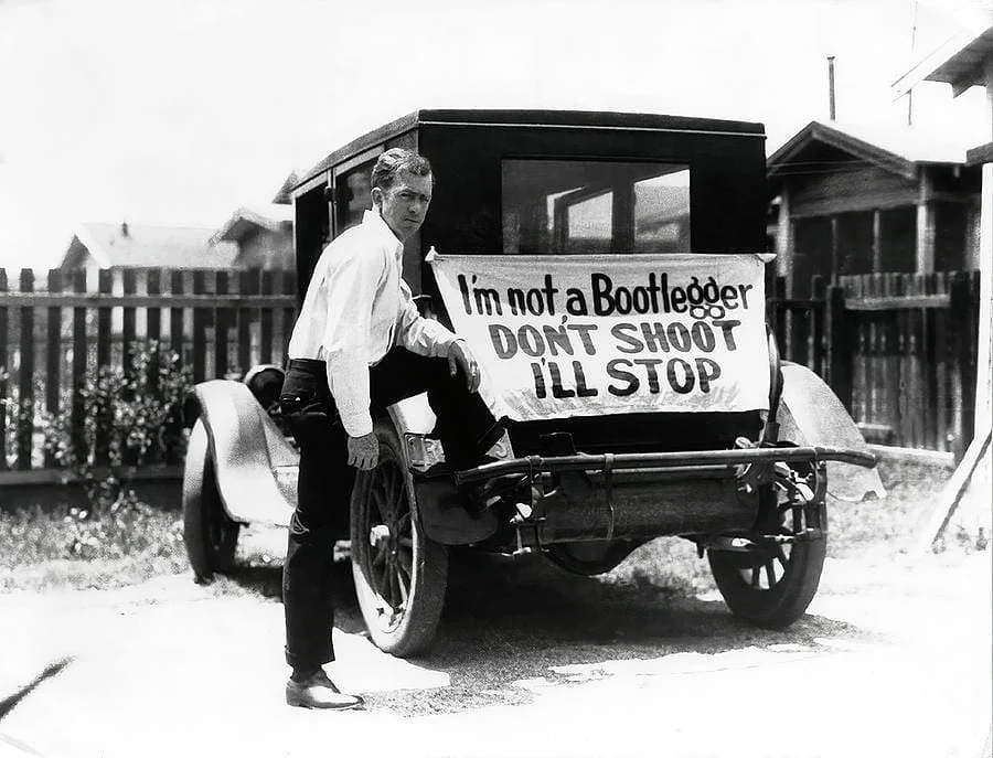 Man standing beside a vintage car with a sign saying "I'm not a Bootlegger, DON'T SHOOT, I'LL STOP" attached to the rear. The scene is outdoors, near wooden fences and houses. The man is wearing a white shirt and pants.