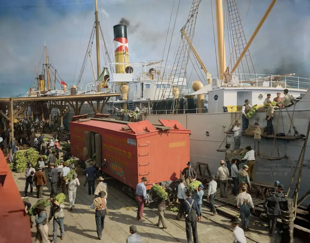 A bustling dock scene with workers unloading bananas from a ship onto a colorful train car. A crowd is gathered, actively moving cargo. The ship has a tall smokestack, and there are cranes and ropes in the background under a partly cloudy sky.