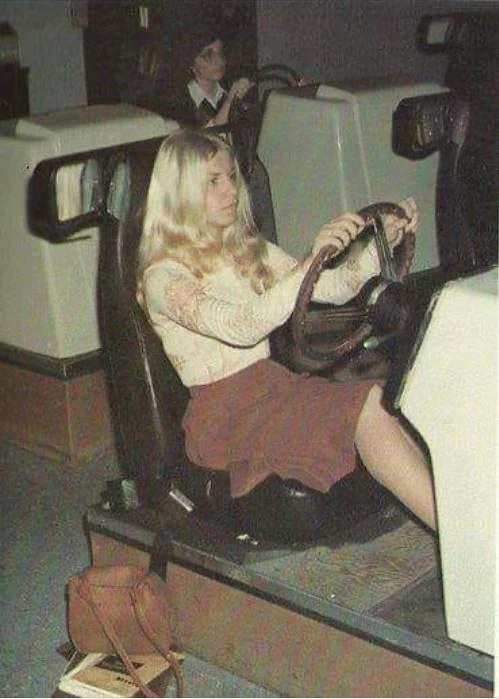 A woman with long blonde hair sits in a vintage arcade driving simulator, focused on the road. She wears a white sweater and a red skirt. A brown handbag is on the floor beside her. Another person is seen in the background also in a simulator.