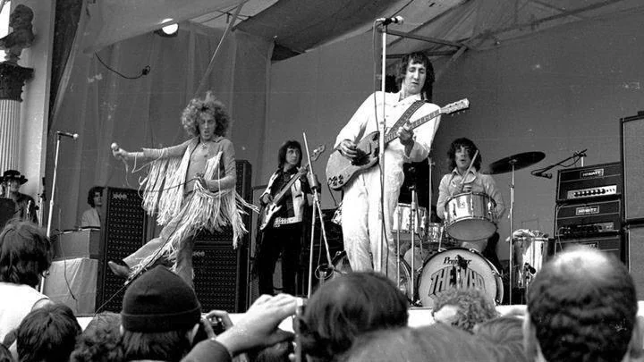 A black and white photo of a rock band performing on stage. The lead singer is wearing a fringed outfit, energetically moving with a microphone. A guitarist and bassist stand nearby, and a drummer is playing. The audience is visible in the foreground.