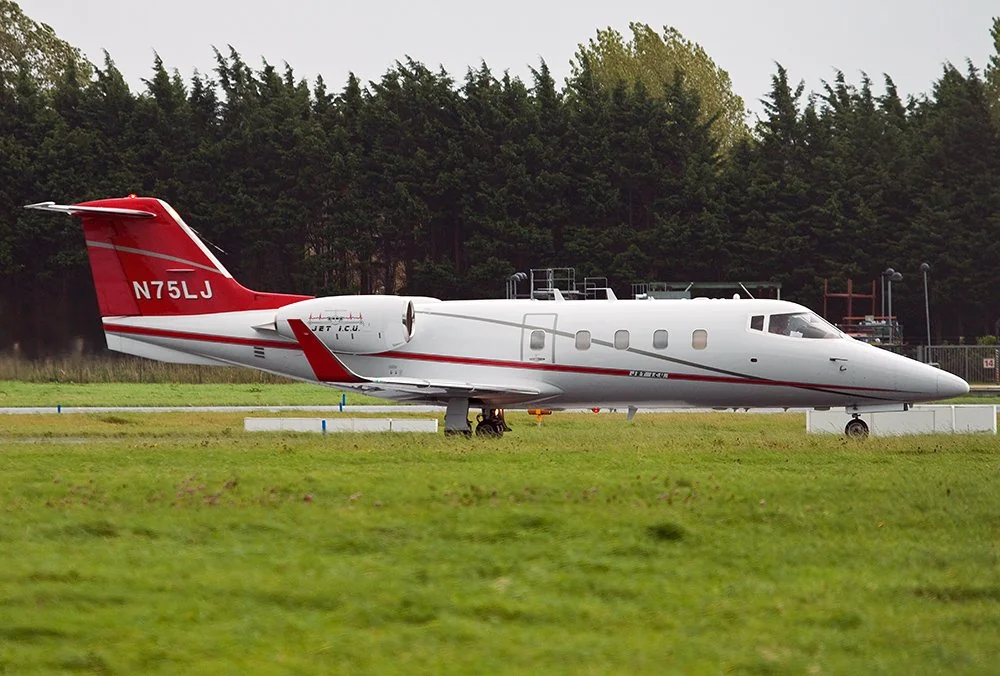 A white and red Learjet 35A on a grassy runway, surrounded by trees. The aircraft has the registration N75LJ visible on the tail. The sky is overcast.