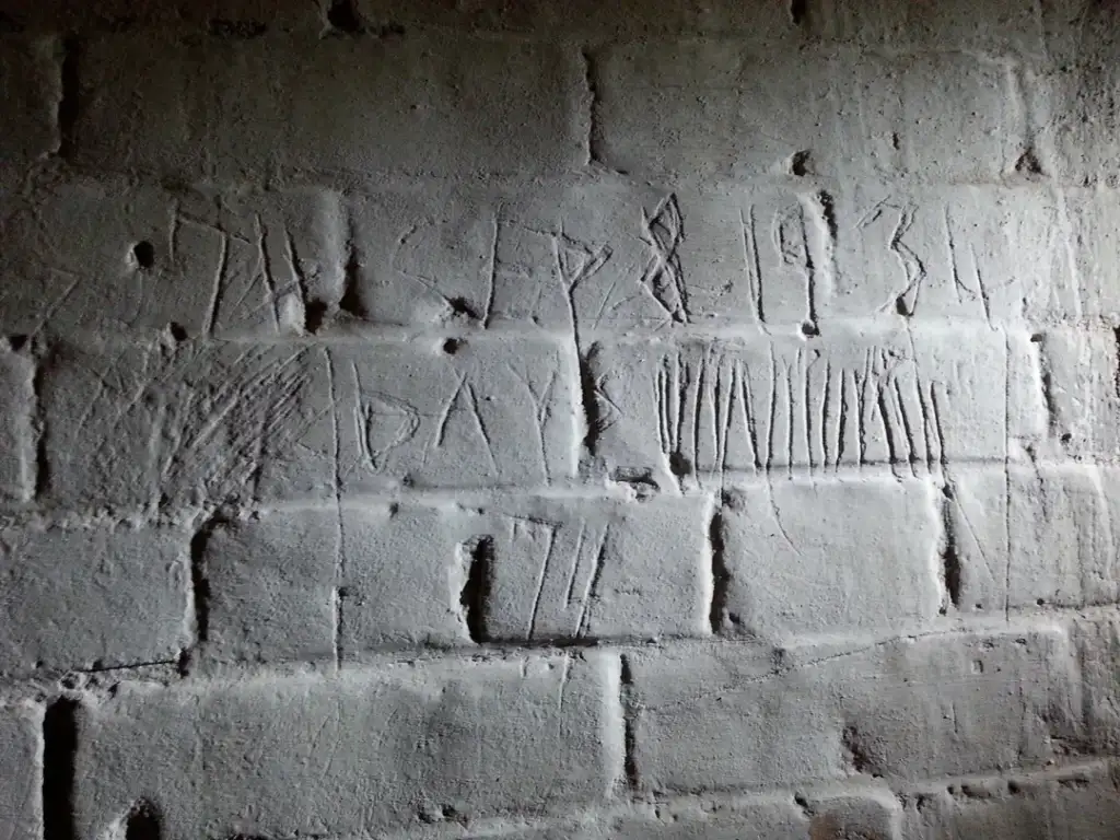 Wall with rough, hand-carved engravings including the date "Sep 8, 1934" and several tally marks. The surface is uneven, with shadows highlighting the texture of the bricks and carvings.