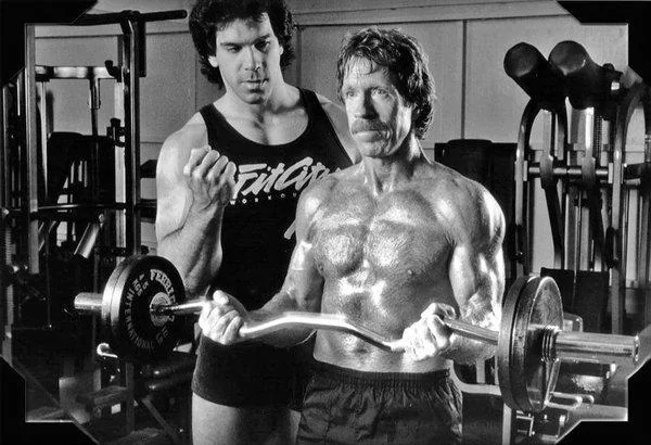 Two men in a gym; one is guiding the other in lifting a weighted barbell. The man lifting is shirtless and muscular, the guide wears a tank top. Gym equipment is visible in the background. The scene is black and white.