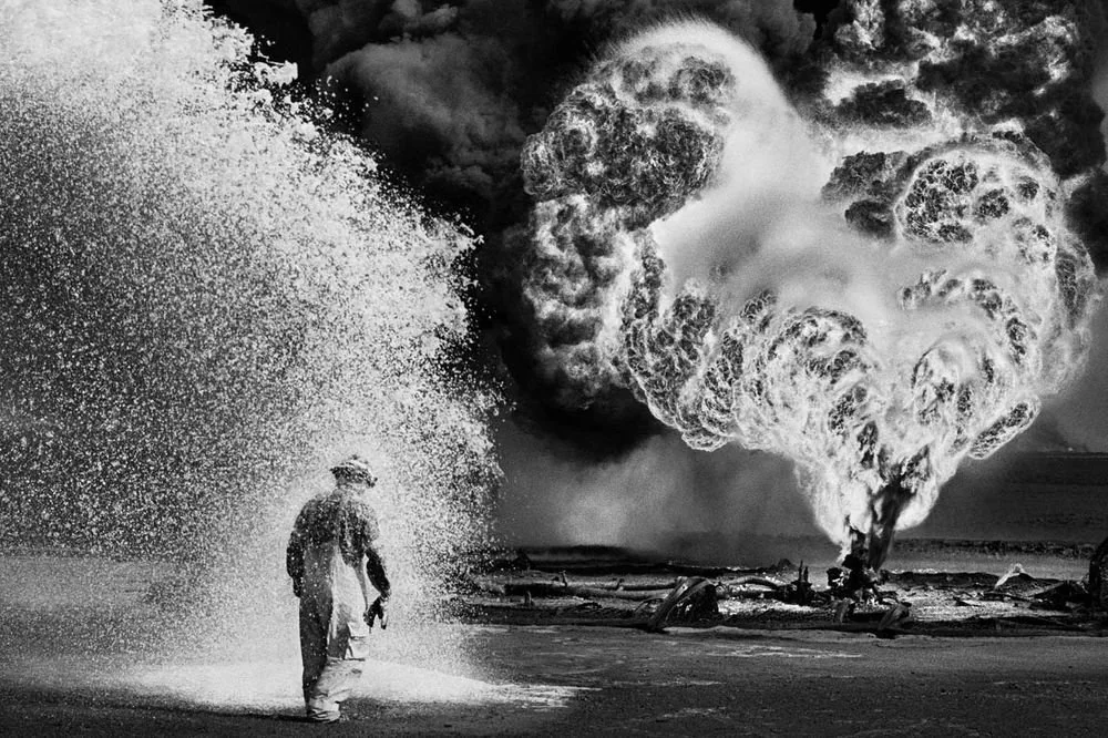 A firefighter in protective gear is engulfed in a cloud of foam as a massive plume of smoke and fire rises dramatically in the background. The scene is intense and action-packed, capturing the dangerous and chaotic nature of the event.