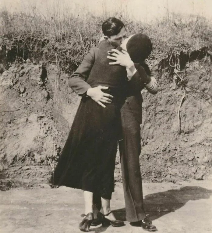 A black and white photo of a couple embracing and kissing. They are standing outdoors against a backdrop of grass and earth. The woman wears a long, dark dress, and the man is in a suit.