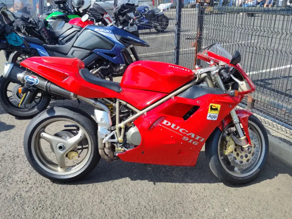 A red Ducati 916 motorcycle is parked on a paved surface, with other motorcycles visible in the background. The bike features a streamlined design, visible exhaust, and a "Ducati 916" label on its body. A chain-link fence can be seen behind it.