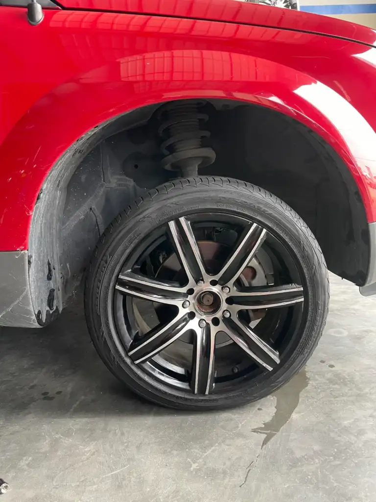 A red car with a close-up view showing a black alloy wheel with a silver detailed rim. The tire is mounted, and the suspension spring is visible above the wheel inside the wheel arch. The car is parked on a concrete surface.