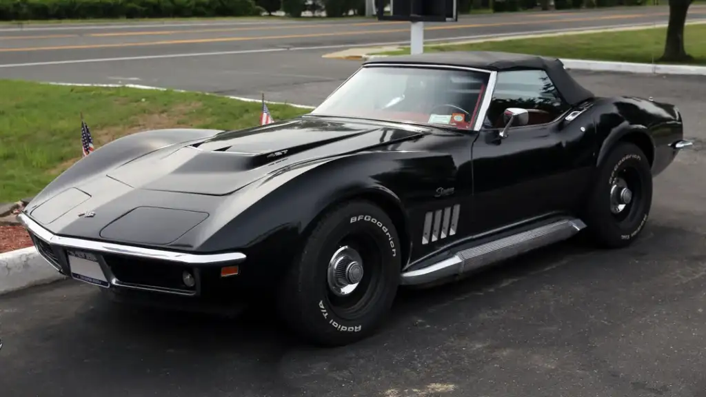 A classic black convertible sports car parked on a paved surface. It features a sleek, aerodynamic design with distinctive front fender vents and a soft top. Small American flags are placed nearby, and a road is visible in the background.