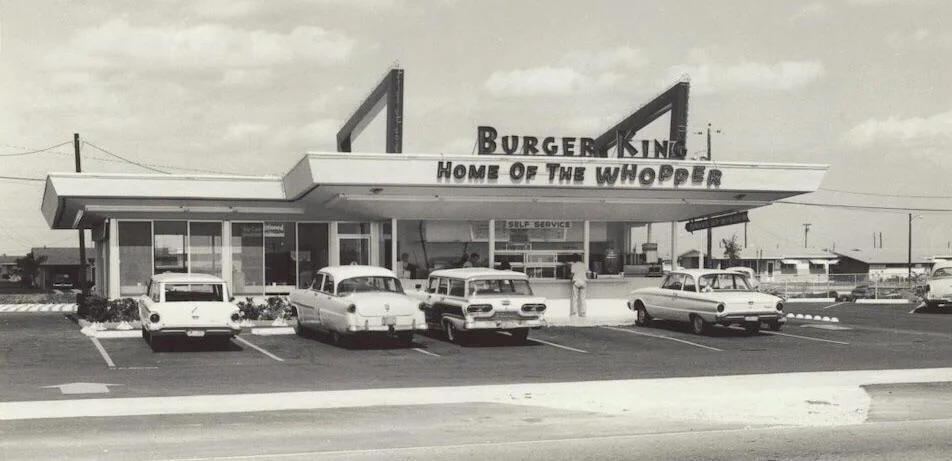 A vintage Burger King restaurant with the sign "Home of the Whopper" is shown. Several classic cars are parked in the lot. The building features retro architecture with large windows and a prominent sign. The setting appears to be a sunny day.