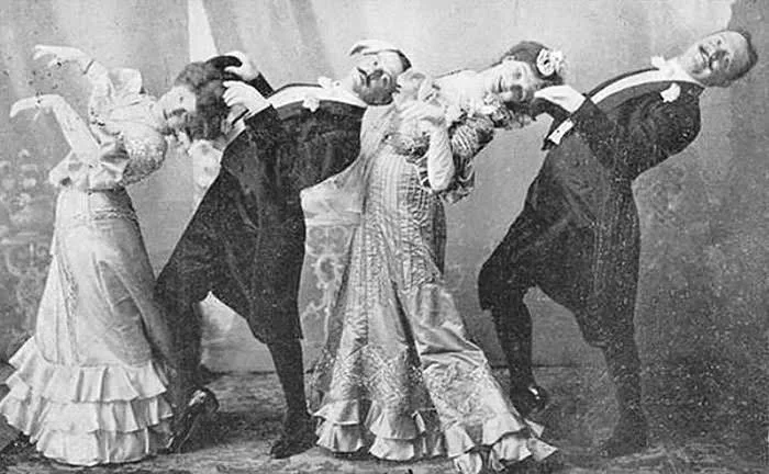 A vintage black and white photo of two women and two men, dressed in formal late 19th or early 20th-century attire, leaning backward in a theatrical pose. The group stands on a patterned carpet with a curtain backdrop.