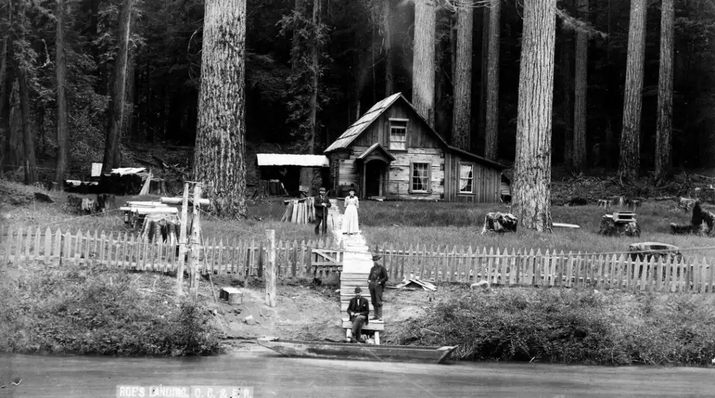 A rustic wooden cabin stands among tall trees with a small picket fence in front. A narrow walkway leads from a canoe by the water to the house. Several people are near the canoe and outside the cabin, surrounded by nature.