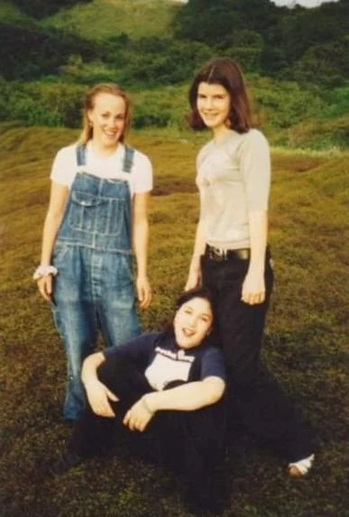 Three young women are outdoors on grassy terrain. One is standing in denim overalls, another in a gray top and black pants, and the third is sitting on the ground in a dark shirt. Lush greenery is visible in the background.