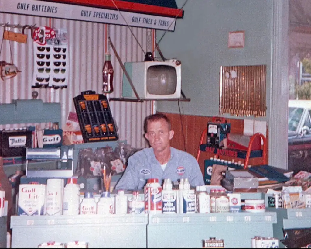 A man in a blue uniform sits behind a counter in a store filled with automotive products. Shelves display oils, batteries, and car accessories. A vintage TV is on the wall, and a window shows a parked car outside. Vintage store decor is visible.